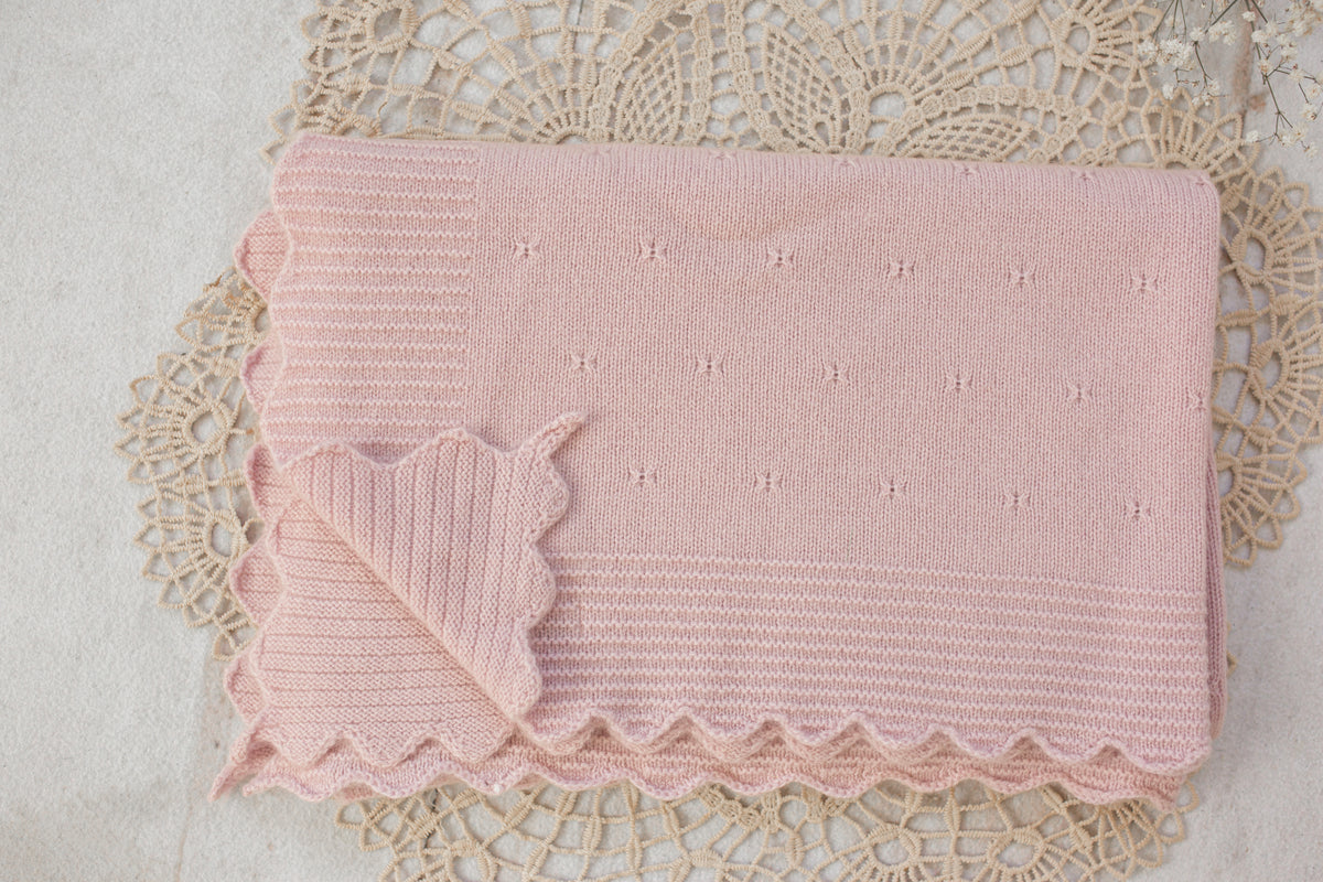 A pale pink knitted baby blanket with a scalloped edge and small embroidered star details, laid flat on a beige lace doily.