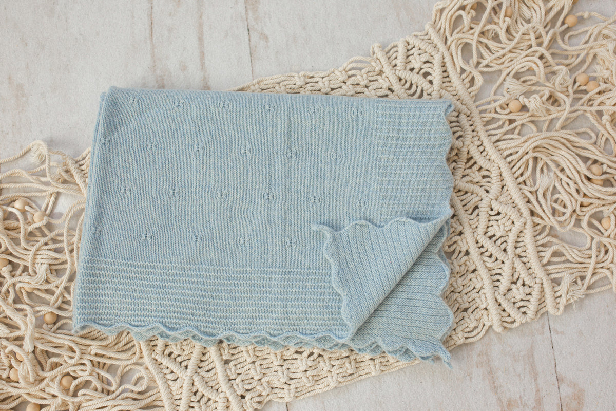 A light blue knitted throw blanket with a scalloped edge, laid flat on a cream-colored macrame runner over a light wood surface.
