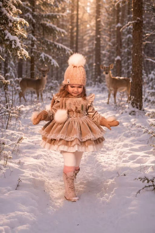 A young girl wearing a beige winter outfit with a matching knit hat and pom-pom boots stands in a snowy forest, surrounded by deer. The outfit includes a layered ruffled dress and a decorative belt.